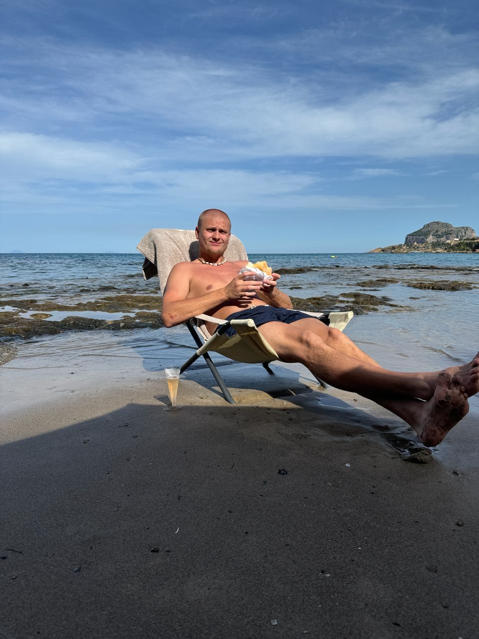 Relaxing in a beach chair at the water's edge on Mazzaforno Beach, Cefal&ugrave;, with the rocky headland in the distance